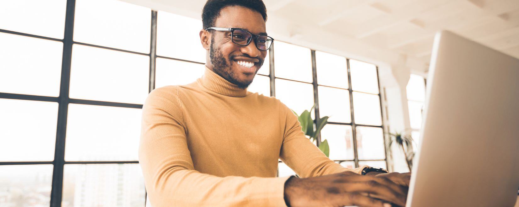 An African-American man with a beard and glasses smiling as he types on a laptop.