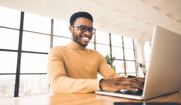 An African-American man with a beard and glasses smiling as he types on a laptop.