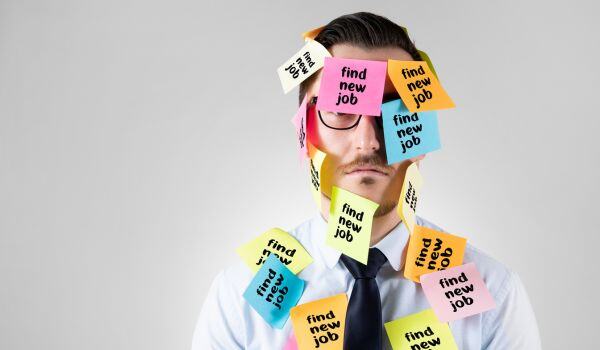 A man with glasses in a shirt and tie with his face covered with post-it notes saying “find new job.”