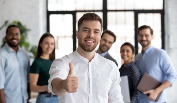 A young man with a beard and dress shirt gives a thumbs-up to the camera while a group of young multiracial professionals smiles behind him. 