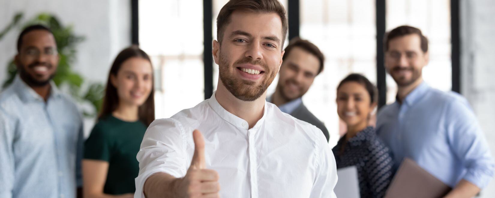 A young man with a beard and dress shirt gives a thumbs-up to the camera while a group of young multiracial professionals smiles behind him. 