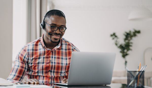 An African-American man wearing glasses and a headset and smiling at a laptop